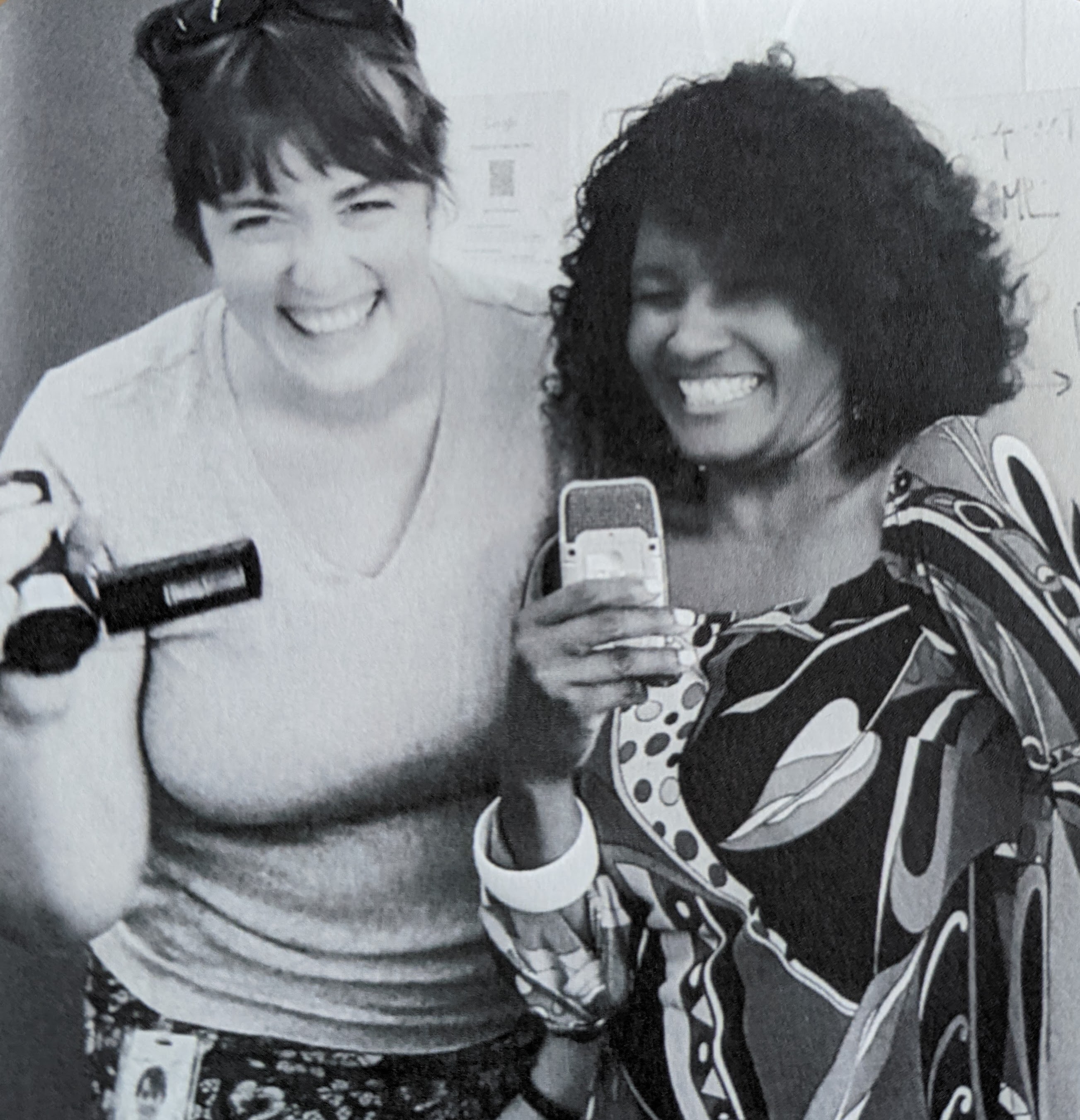 Two women are in the foreground of this black and white picture, Katie, a tall white woman with short dark hair, and Keita, a shorter Black woman with curly shoulder length hair and a fabulously patterned dress. Both women are laughing. Katie is on the left and holds a small camcorder. Keita is on the right and holds a small microphone.