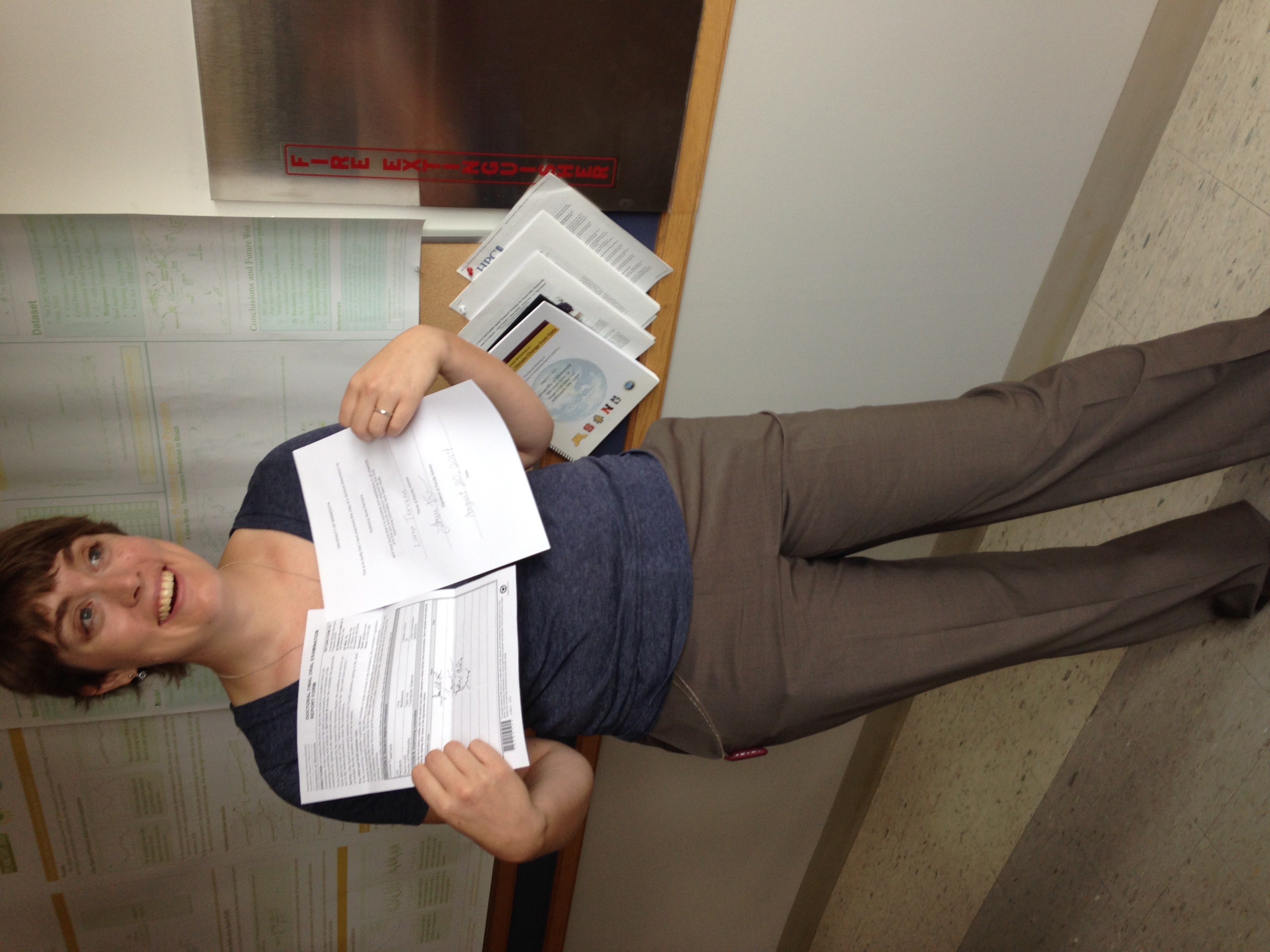 Katie, a white woman with short brown hair, is looking to the right of the photographer. She is wearing a necklace, dark blue shirt, and brown dress pants. In her hands, covering her chest are two pieces of paper. Both have three or four signatures on them. Katie has a goofy smile on her face. There is a corkboard with scientific posters on it in the background and she is standing in a university hallway next to a fire extinguisher box.