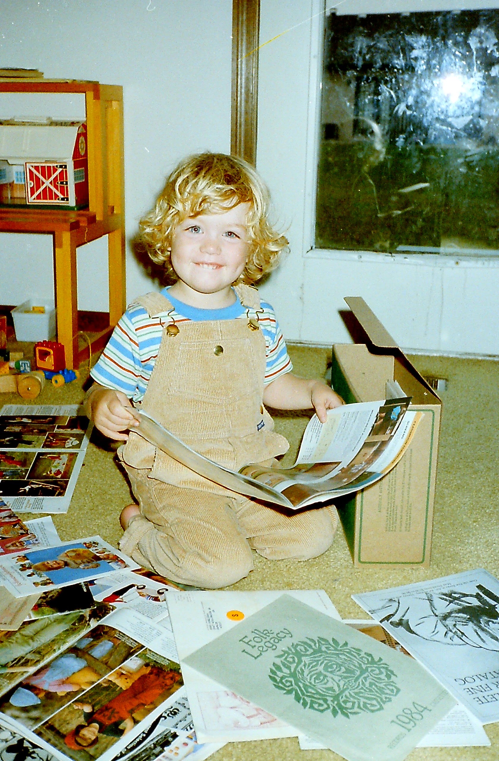 Katie is shown as a wide eyed, curly blonde haired, white toddler, around the age of 3. She is biting her lip and holding a knitting pattern. More colorful patterns are scattered in front of her on the floor and a shelf of toys is in the background.