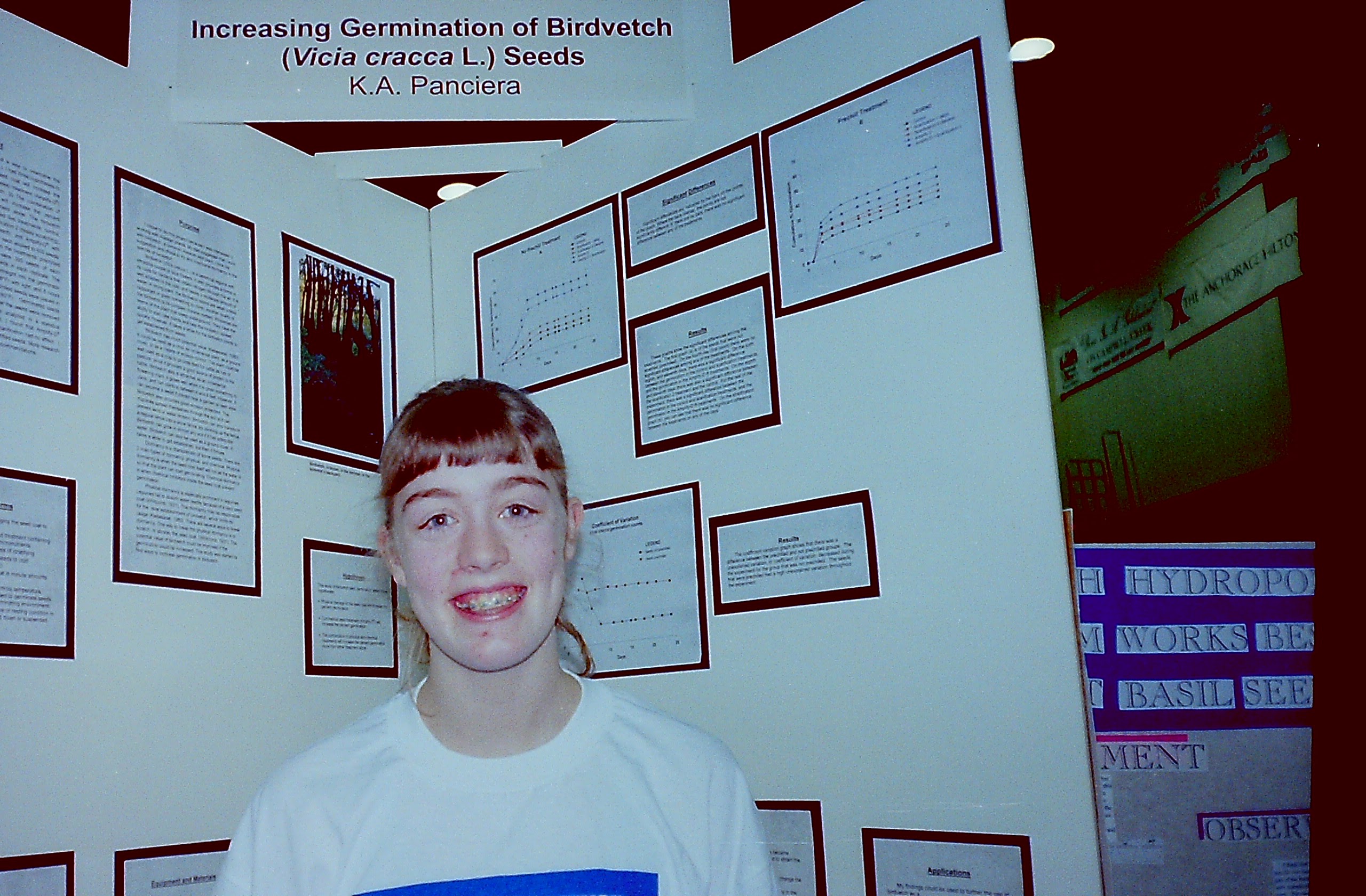 Katie, a white girl with brown hair, bangs, and curls escaping from a ponytail, smiles largely showing off braces, in the foreground. Behind her is a science fair poster. The title at the top is Increasing Germination of Birdvetch (Vicia cracca L.) Seeds K.A. Panciera. The rest of the poster is mainly illegible black text printed on white paper and mounted on black backgroun. On the right above her head is a picture of bird vetch. To her left are several line graphs.