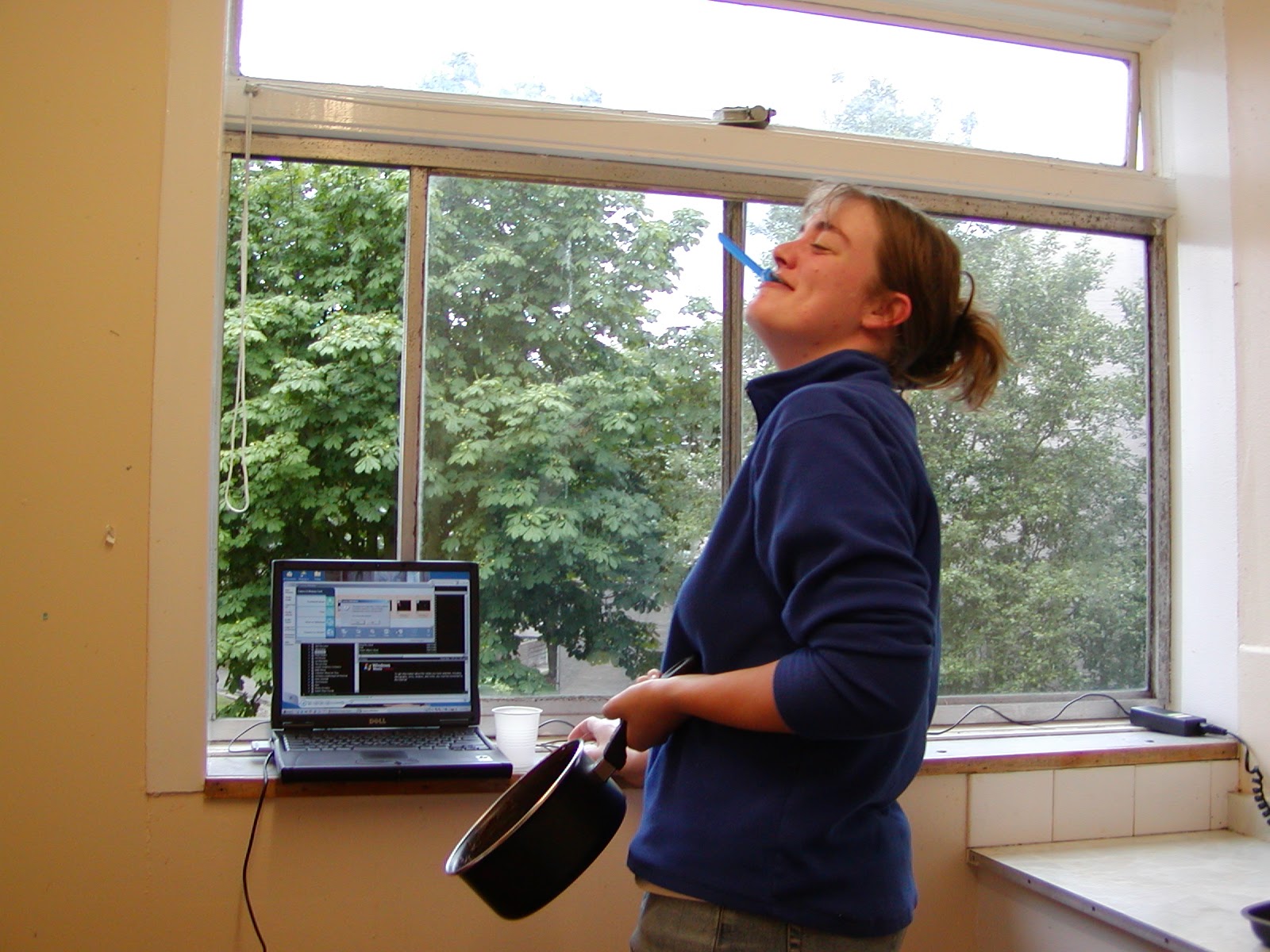 Katie, a white female with brown hair up in a messy clip, is shown in profile looking towards the left side of the picture wearing a blue fleece and jeans. She is laughing with her head tilted up and a blue spoon in her mouth. In her left hand is a pot. She is in a kitchen and there is a large window behind her. There is a view of trees from the window. On the windowsill is a 2003 Dell laptop playing music. The powercord goes across the frame to an unshown outlet.