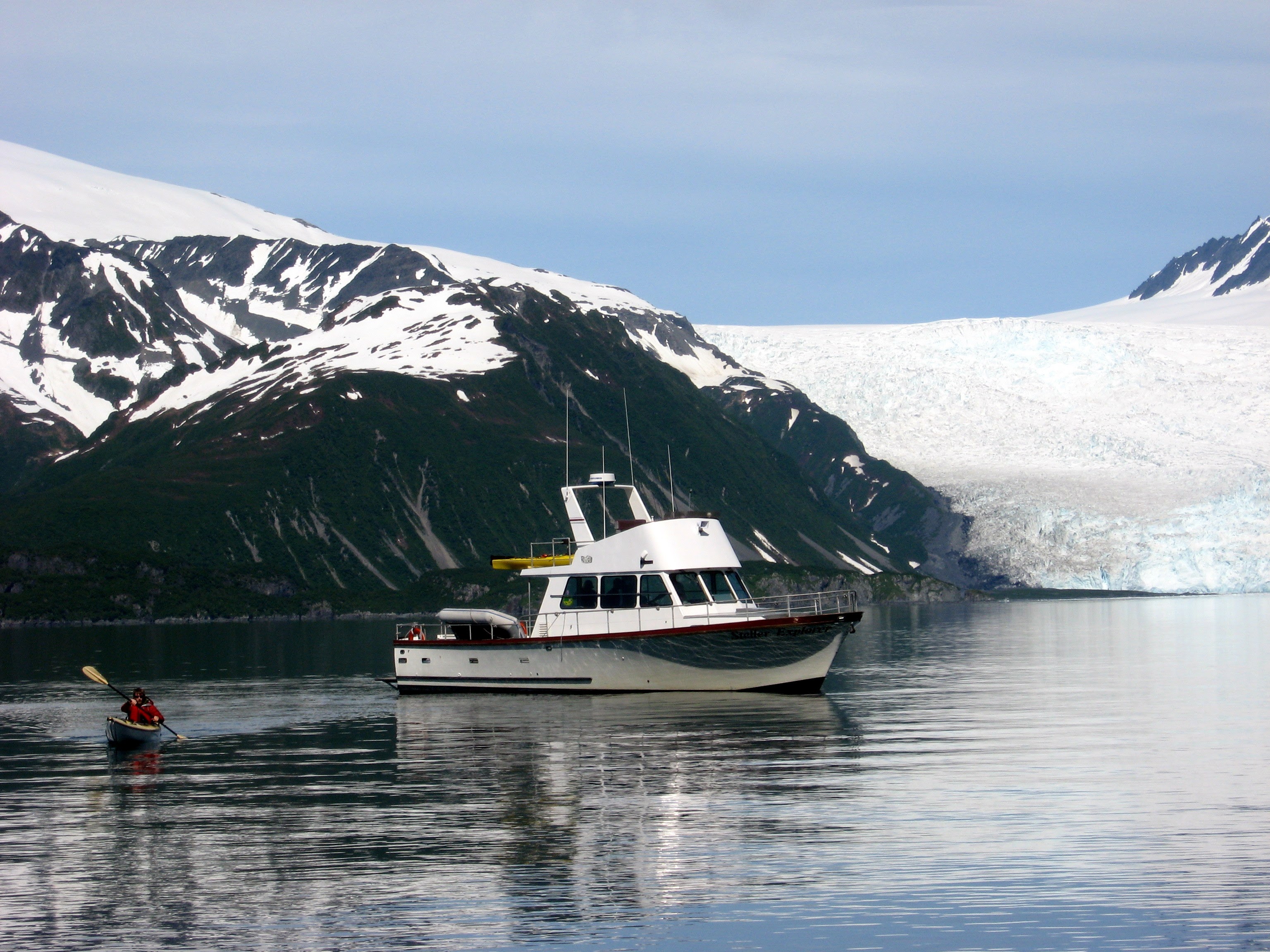 The foreground is a black ocean. On the left a person is kayaking in a dark kayak with a red vest and tan oars. To their right is a small white sightseeing boat. There is comfortable room for 6-8 people on the boat in nice weather. There is a yellow kayak on the roof of the cabin and a gray raft behind the cabin. In the background on the right is a white glacier with occasional tints of gray and light blue. It is touching the water. On the left, there is a dark mountain with a number of avalanche trails. The mountain is covered with snow at the top and the snow continues til about halfway to the water. 