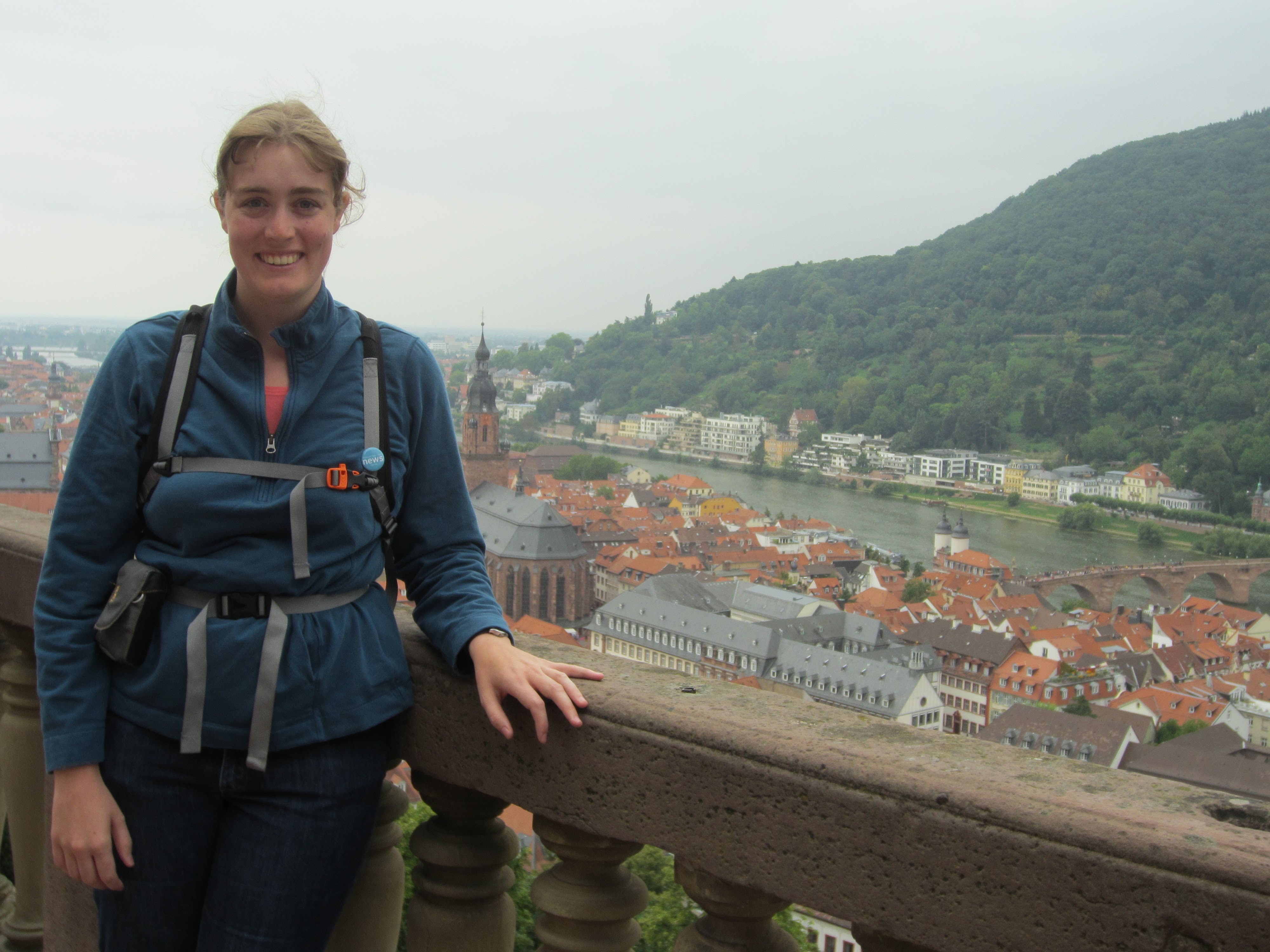 Katie, a white woman, with pulled back brown hair stands on the left, pictured from wais up. She is wearing a teal quarter zip fleece, unzipped with a coral shirt undernearth. Her gray backpack straps are very visible including a waistbelt and chest strap. The chest strap buckle is neon orange. There is a MPR news button on her right backpack strap. She is leanding casually on a stone wall. Behind her you see the primarily red rooves of the old neighborhoods of Heidelberg. Behind them is a river, the Neckar river, with bridge on the far right. On the other side of the river is one strip of more modern buildings and behind that is a green wooded mountain. The Heiliggeistkirche appears to the right of Katie's elbow.